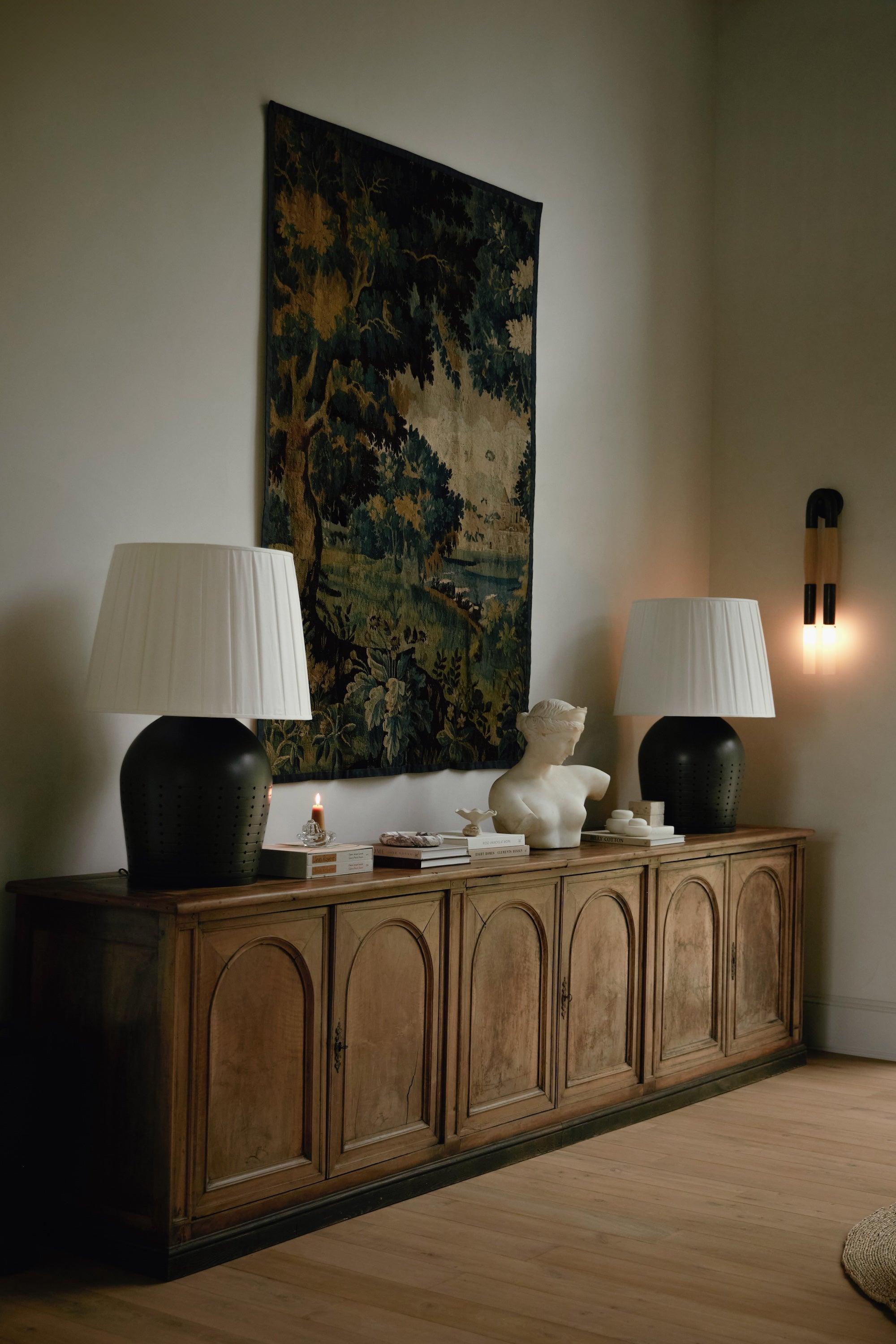 An image of a wooden credenza with two large table lamps and a tapestry hanging on the wall.