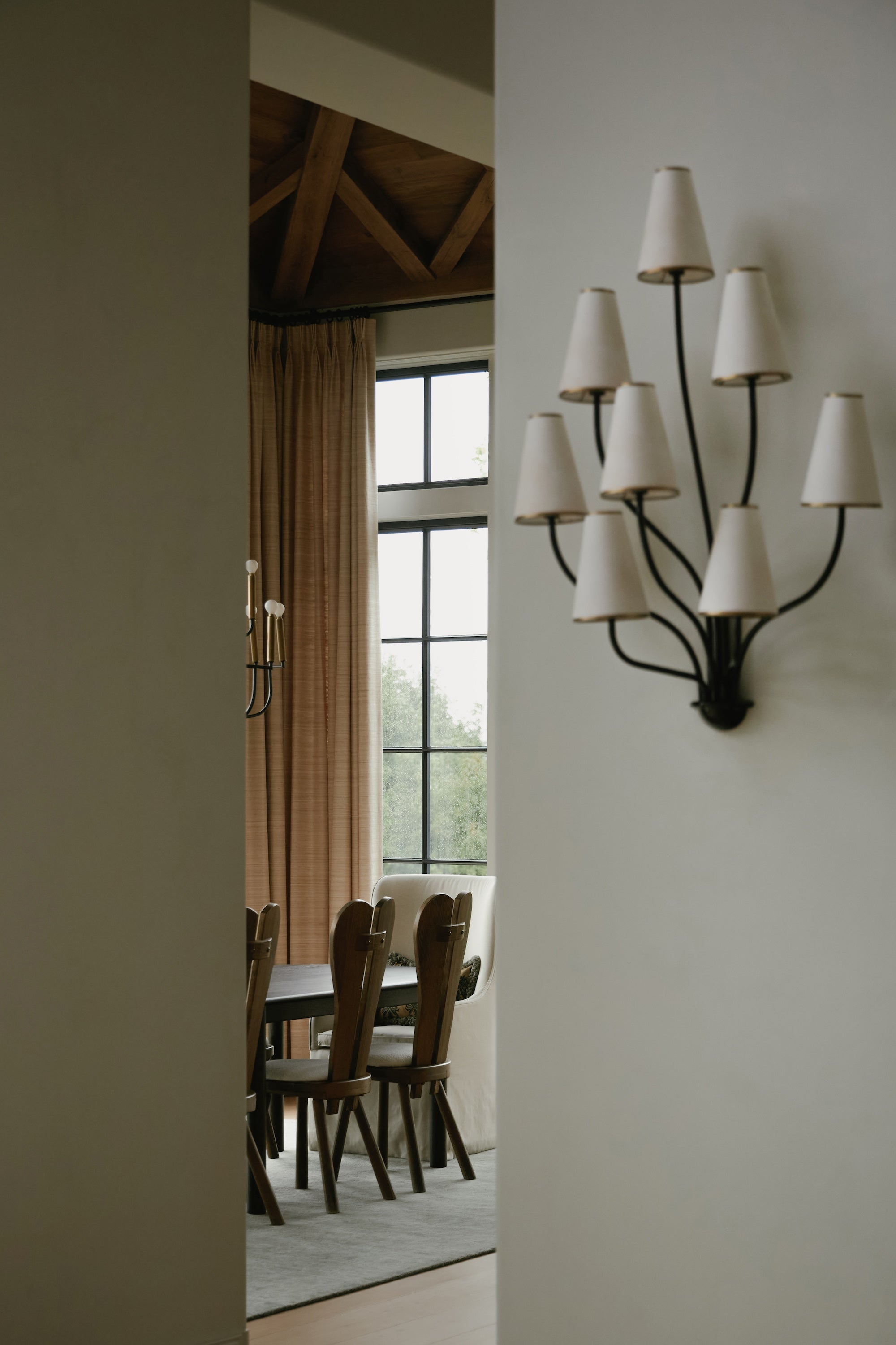 An image looking into a dining room, featuring a wall sconce with black metal arms and fabric shades.